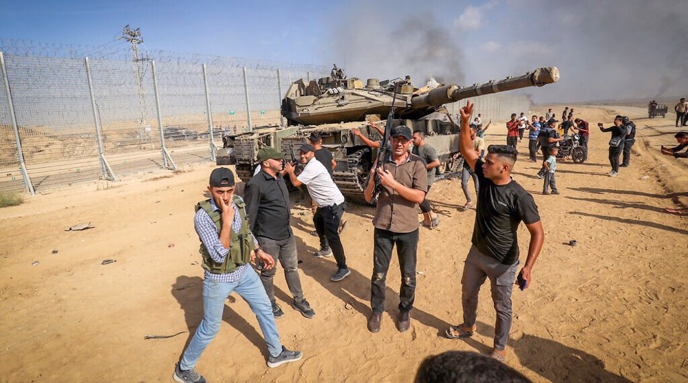 فلسطينيون يرفعون شارات النصر أمام دبابة إسرائيلية بمحاذاة السياج الحدودي مع غزة، وذلك بعد الاختراقات التي أحدثتها المقاومة في ذلك السياج، في اليوم الأول لمعركة طوفان الأقصى.