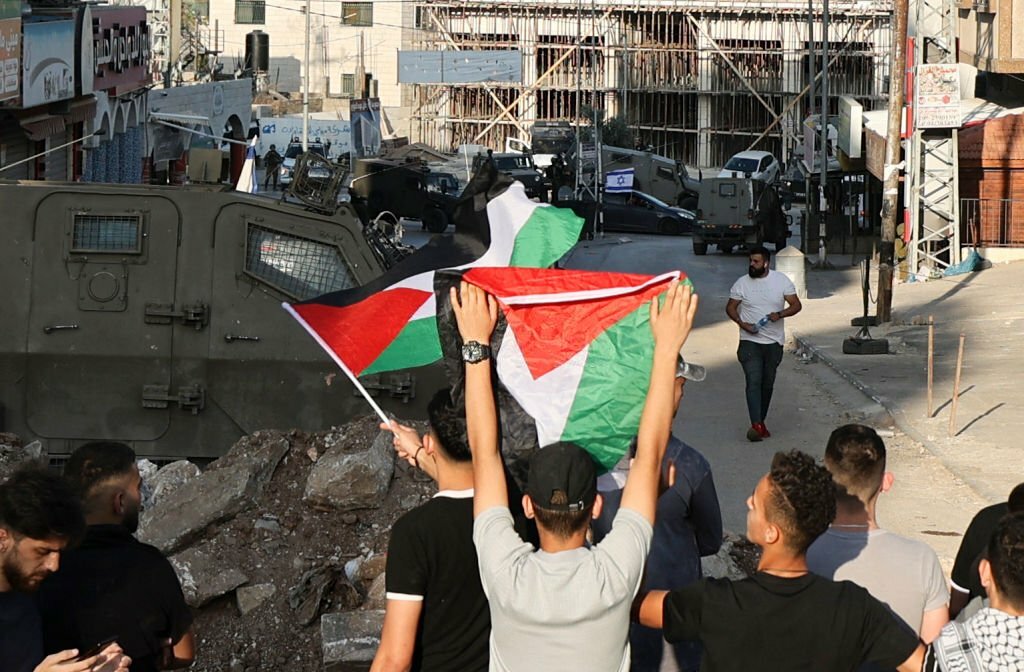 جيش الاحتلال يغلق طريقاً ترتبط بشارع حوارة الرئيس، بينما يرفع فلسطينيون العلم الفلسطينيّ في مواجهة مستوطنين يرفعون علم الاحتلال، أيار/مايو 2022. (عدسة: جعفر اشتية/ وكالة الصحافة الفرنسية).