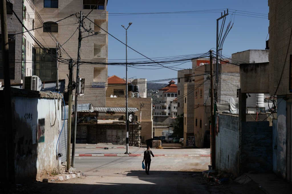 حوارة، الضفّة الغربيّة، تشرين الثاني/ نوفمبر 2023، شاب فلسطيني يمر بالقرب من شارع حوّارة الرئيس، بينما تظهر محلات تجارية وقد أجبرها الاحتلال على إغلاق أبوابها منذ معركة طوفان الأقصى. (Getty Images).